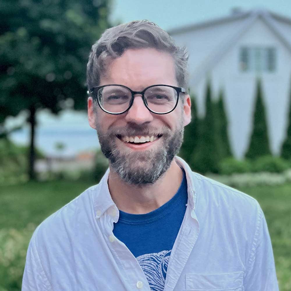 Headshot of Alex Barron, a bearded man with salt and pepper hair, large black-rimmed glasses, and a bright smile, standing outside with green grass and trees behind him