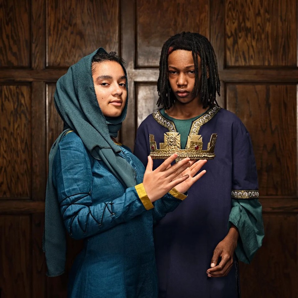 Two teenagers in dark blue historically-inspired Elizabethan costumes stand in front of a deep brown wood wall. The teen in front, dressed in a kirtle, stares at the camera while holding a crown.